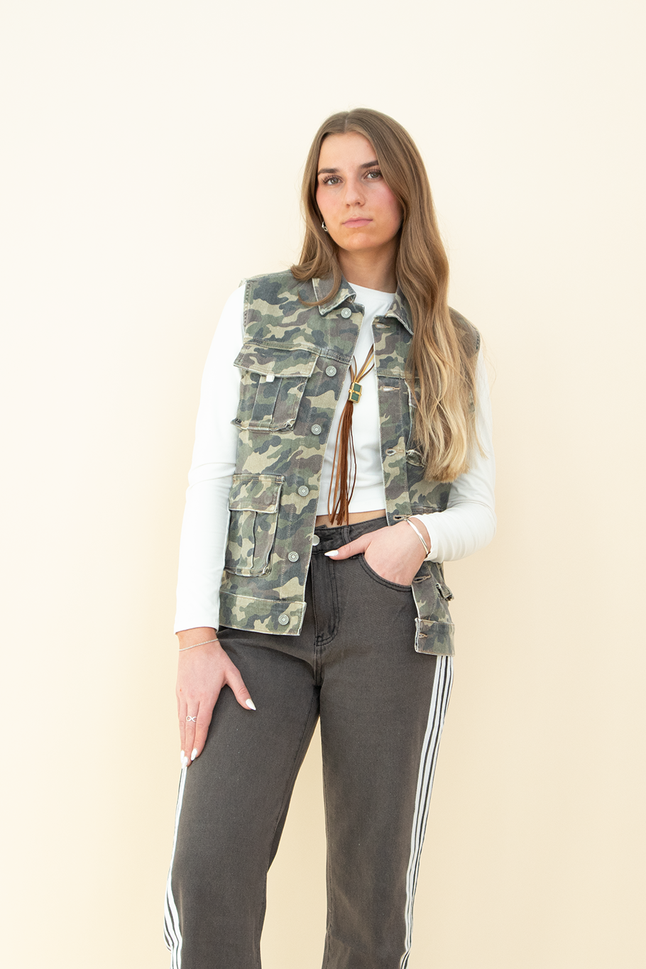 Woman wearing a sleeveless camouflage utility vest with multiple front pockets, styled over a long-sleeve white crop top and paired with charcoal gray track pants with white side stripes. Trendy camo vest for women, perfect for streetwear or casual layering.