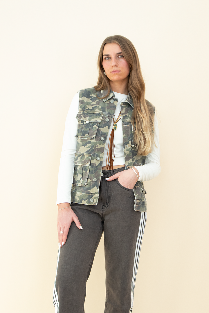 Woman wearing a sleeveless camouflage utility vest with multiple front pockets, styled over a long-sleeve white crop top and paired with charcoal gray track pants with white side stripes. Trendy camo vest for women, perfect for streetwear or casual layering.