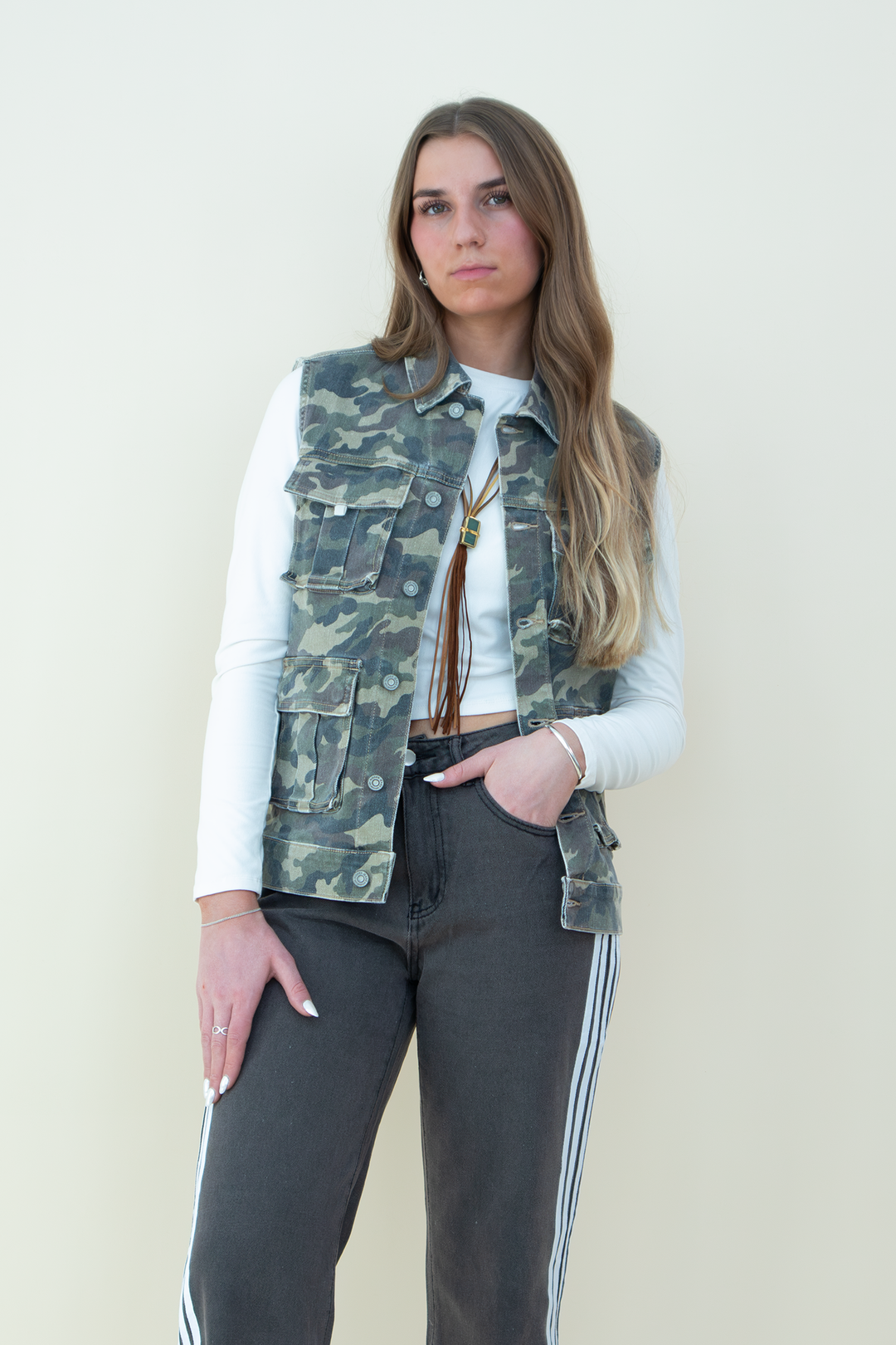 Woman wearing a sleeveless camouflage utility vest with multiple front pockets, styled over a long-sleeve white crop top and paired with charcoal gray track pants with white side stripes. Trendy camo vest for women, perfect for streetwear or casual layering.