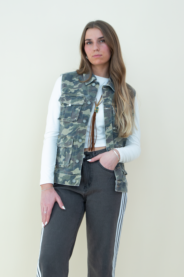Woman wearing a sleeveless camouflage utility vest with multiple front pockets, styled over a long-sleeve white crop top and paired with charcoal gray track pants with white side stripes. Trendy camo vest for women, perfect for streetwear or casual layering.