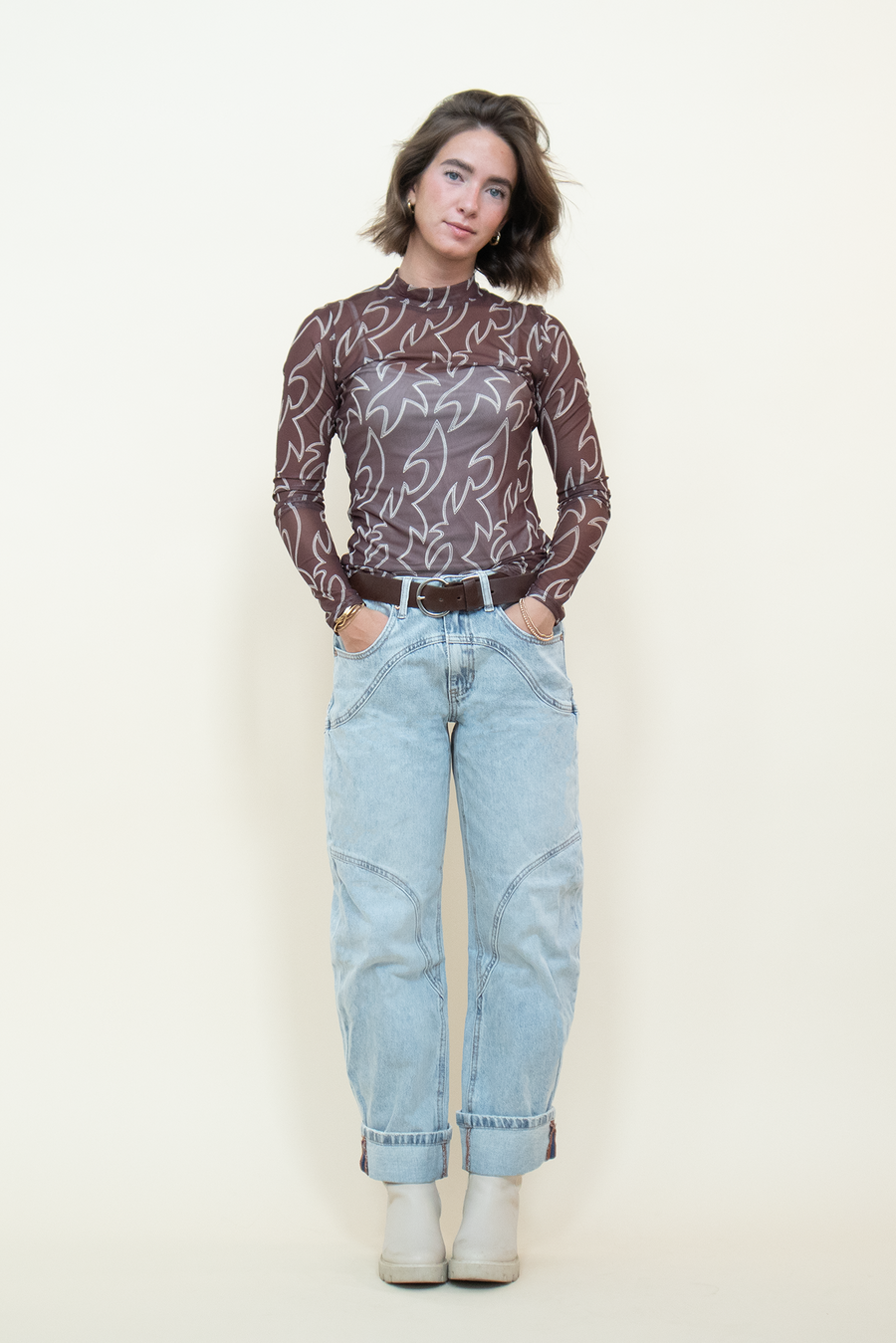 Model wearing a brown mesh top, cream boots and light denim jeans with seam detailing in front of a beige backdrop.