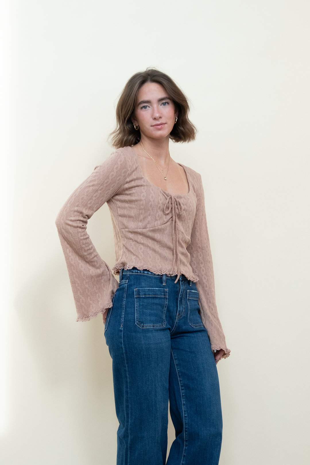 Dusty pink long sleeve crop top with square neckline, front tie bow detail, bell sleeves, and lettuce lace hem, styled with wide leg blue jeans