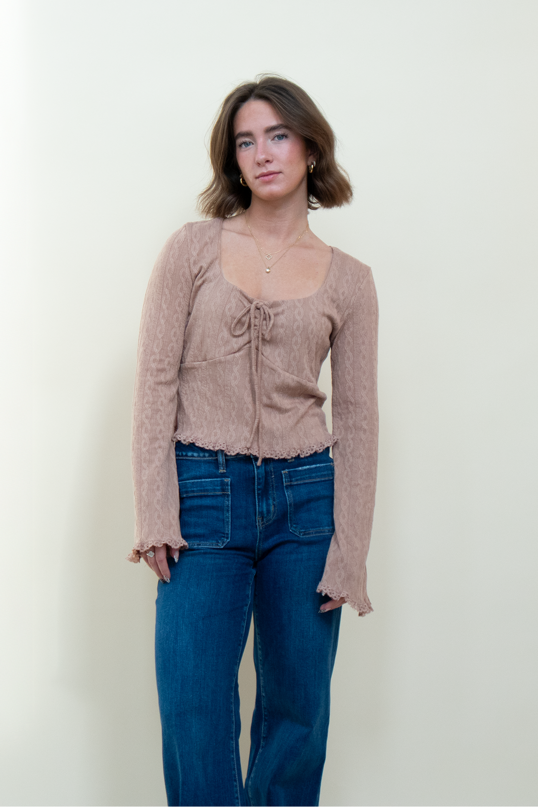 Dusty pink textured long sleeve crop top with square neckline, front tie closure, underbust seam detail, and bell sleeves with lace trim, worn with wide leg jeans