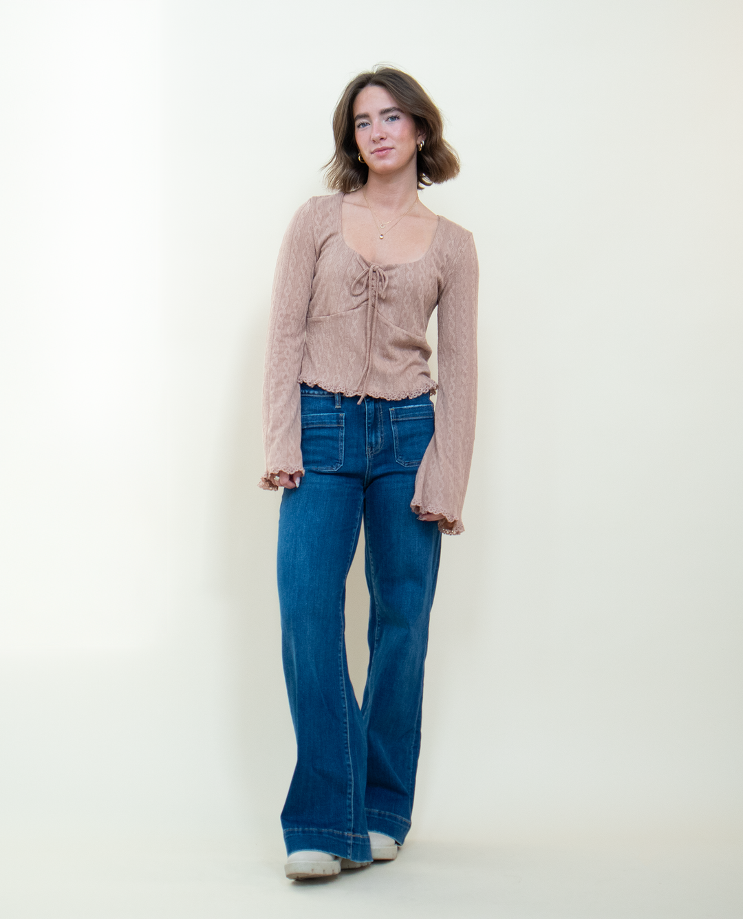 Full length view of dusty pink bell sleeve crop top with front tie and lace hem detail, styled with wide leg blue jeans and white platform shoes