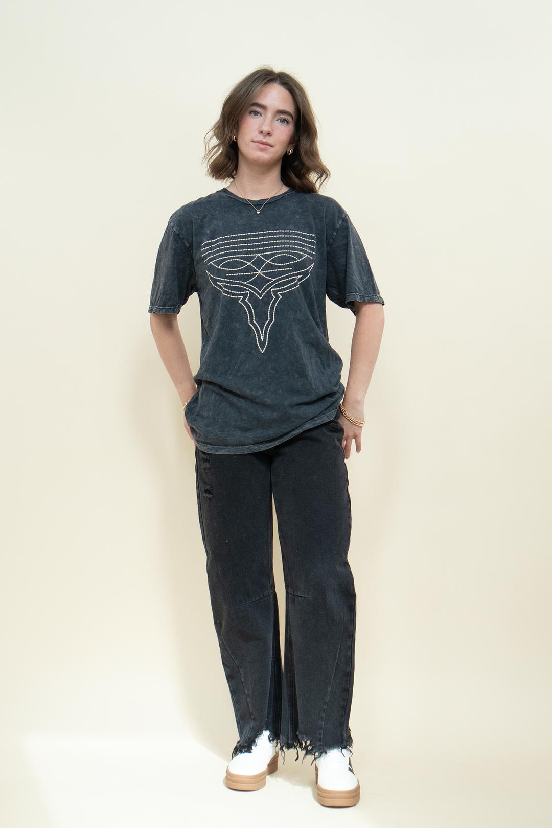 A woman modeling a washed black graphic t-shirt with a white stylized ox design, paired with dark distressed jeans and white sneakers, standing against a solid cream background.