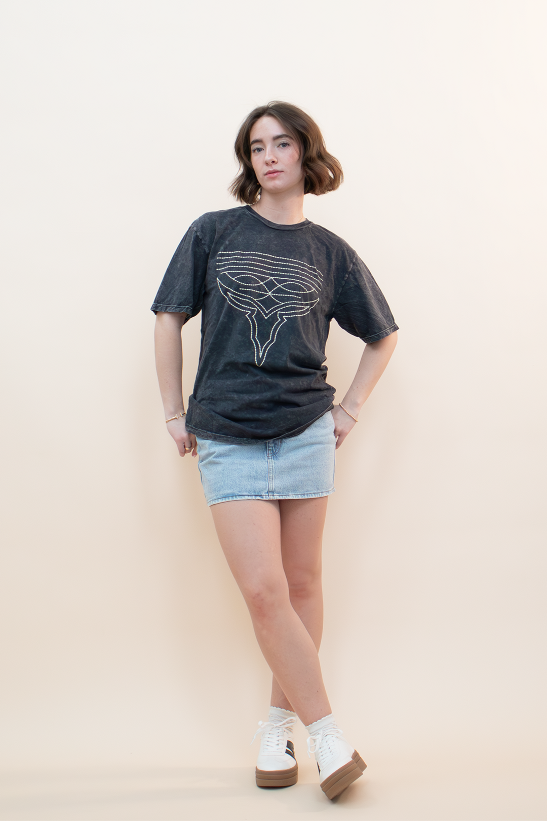 Women’s vintage-inspired graphic t-shirt in washed black with a relaxed fit, paired with a light-wash denim mini skirt. Boutique-style outfit for women 18-40 featuring effortless, everyday style with a touch of edge—perfect for casual chic looks.