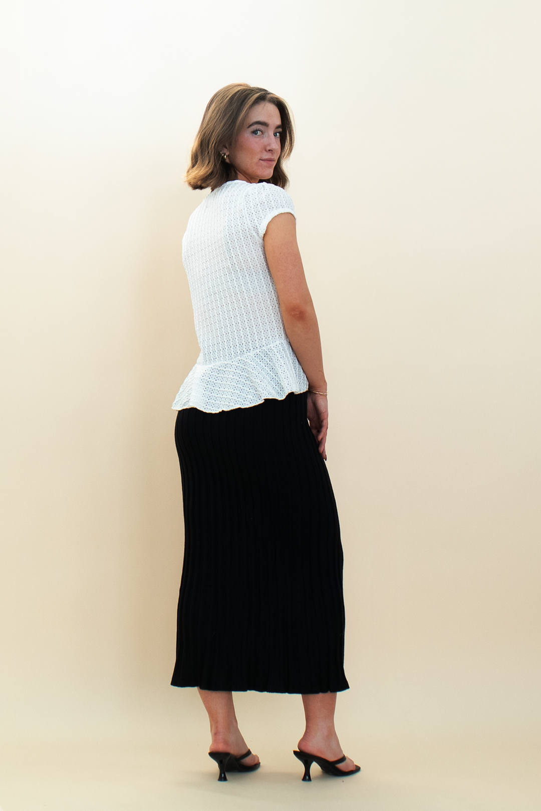 Back view of woman wearing a white textured peplum blouse and black ribbed knit midi skirt, styled with black kitten heels from Groove Closet.
