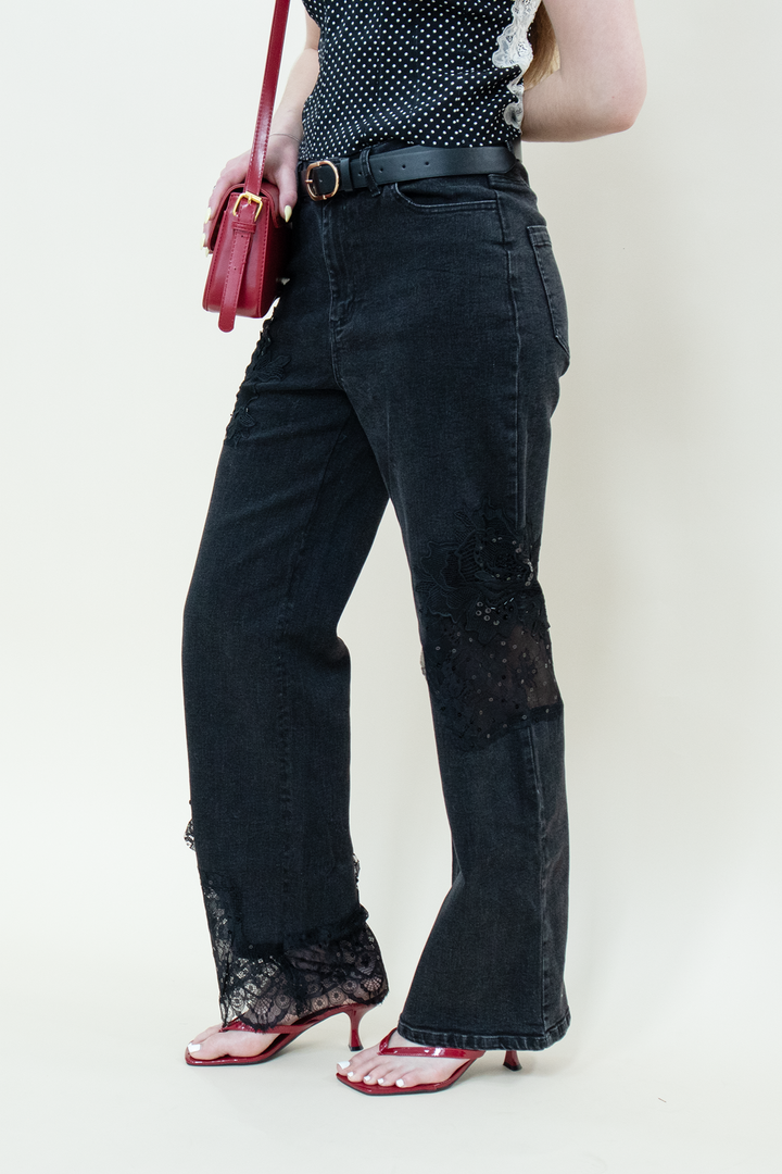 Close-up of a woman wearing black lace-paneled straight-leg jeans from Groove Closet, styled with a black polka dot lace-trim top, red strappy heels, and a matching red handbag.