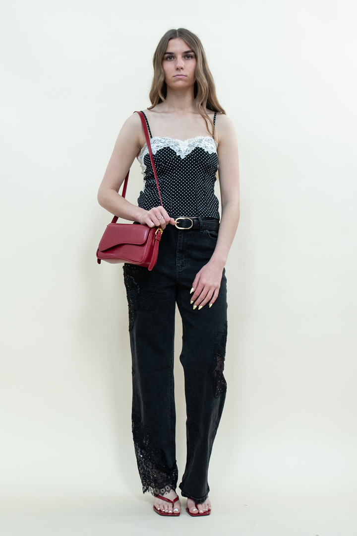 Model wearing black straight-leg jeans with floral lace panels from Groove Closet, styled with a black polka dot camisole with white lace trim, a black belt, red shoulder bag, and red kitten heels.