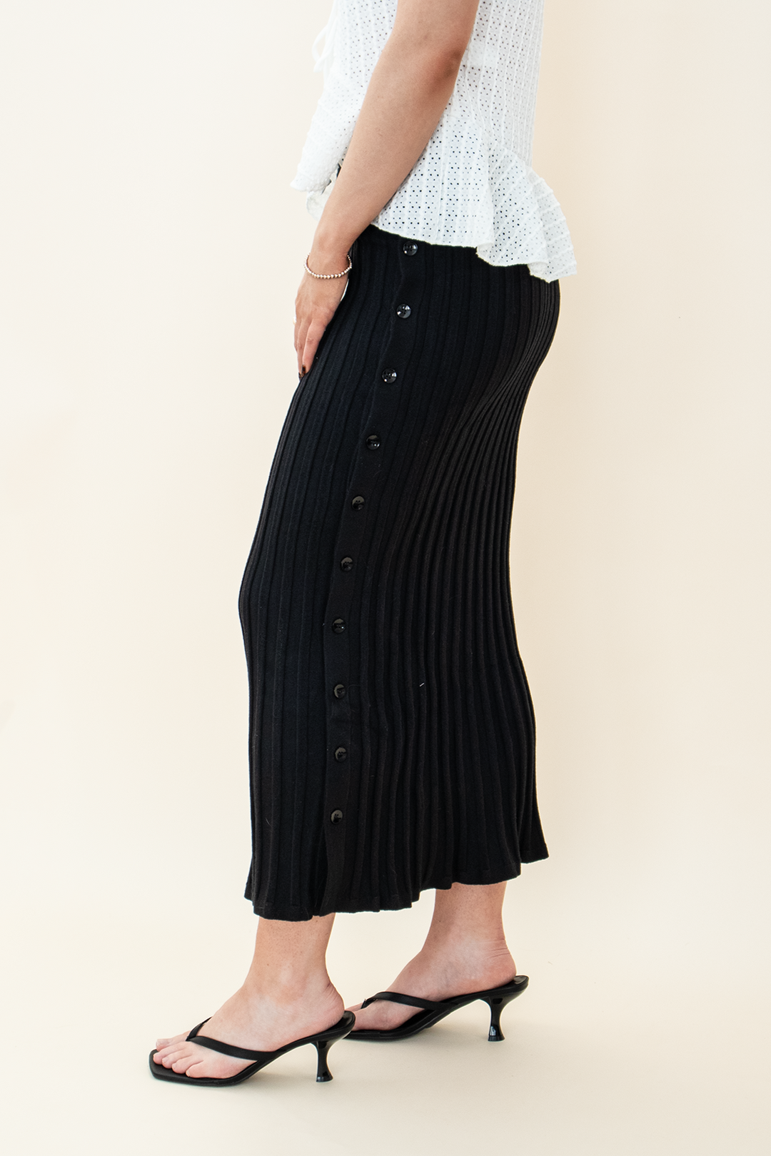 Close-up side view of a black ribbed knit midi skirt with front buttons, paired with black kitten heel sandals and a white peplum top – Groove Closet women’s fashion.