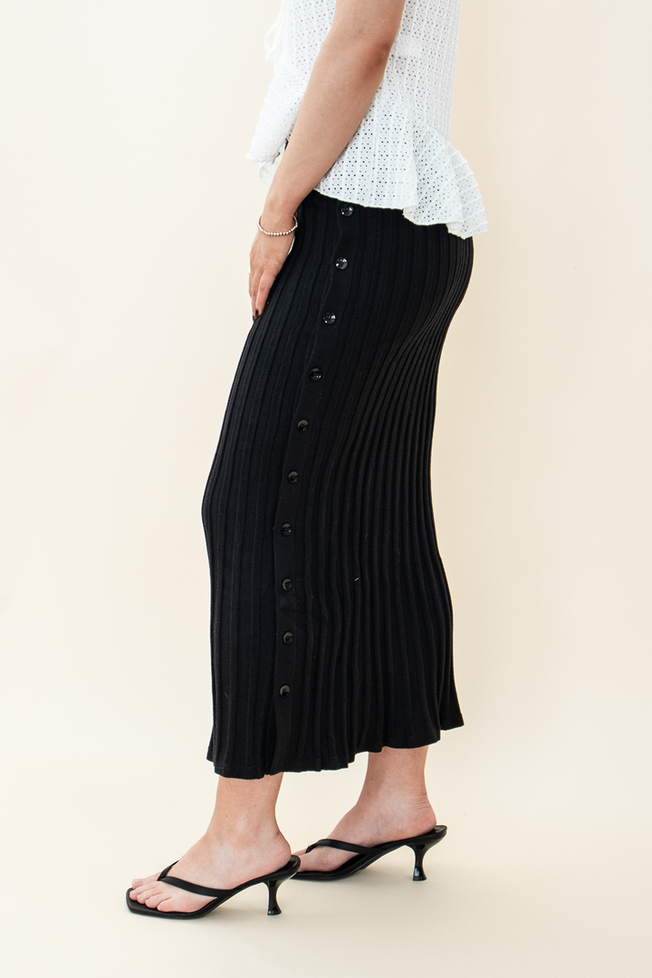 Close-up side view of a black ribbed knit midi skirt with front buttons, paired with black kitten heel sandals and a white peplum top – Groove Closet women’s fashion.