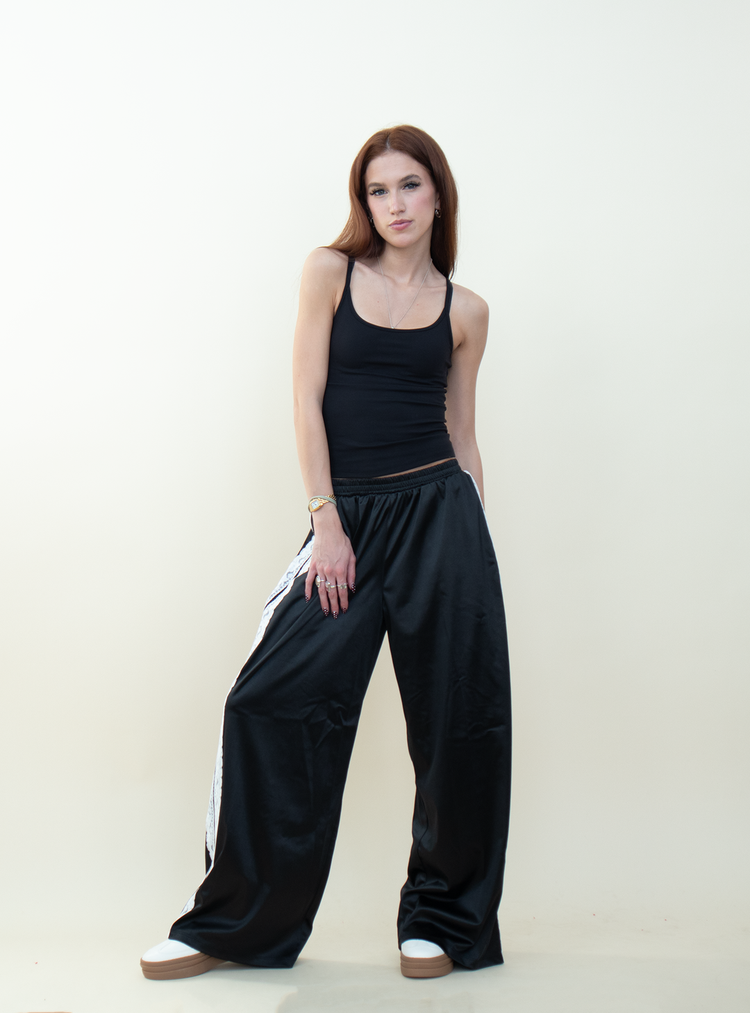 Model wearing black satin wide-leg pants with white lace side stripes, paired with a fitted black tank top and white platform sneakers.