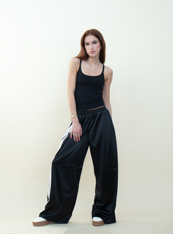 Model wearing black satin wide-leg pants with white lace side stripes, paired with a fitted black tank top and white platform sneakers.