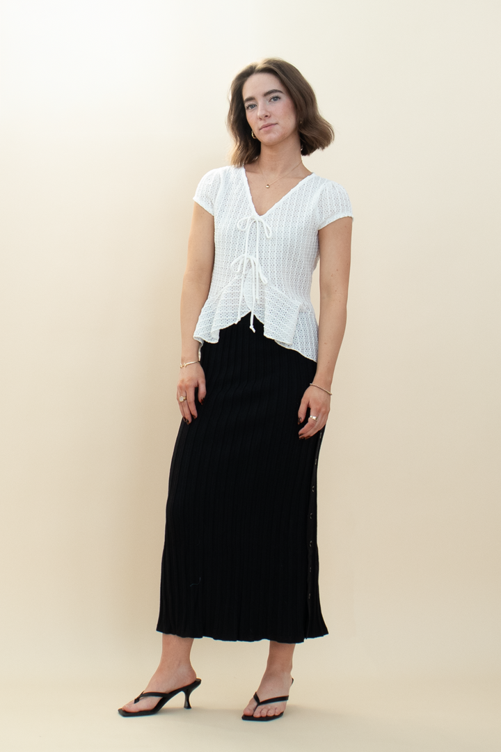 Model in minimalist elegant outfit featuring a white short-sleeve tie-front blouse and a black ribbed midi skirt with slit, styled with black heeled sandals
