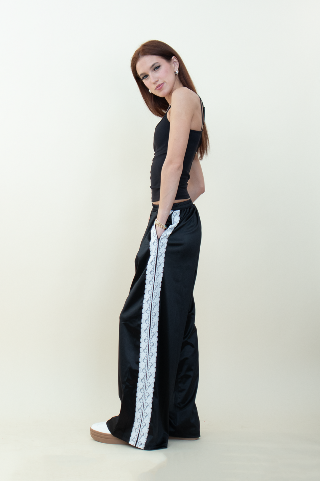 Model standing sideways in black wide-leg pants with lace stripe detail, hands in pockets, styled with a sleek black camisole.
