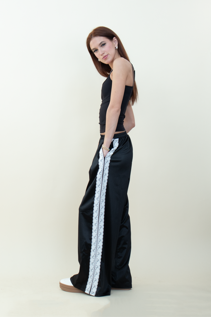 Model standing sideways in black wide-leg pants with lace stripe detail, hands in pockets, styled with a sleek black camisole.