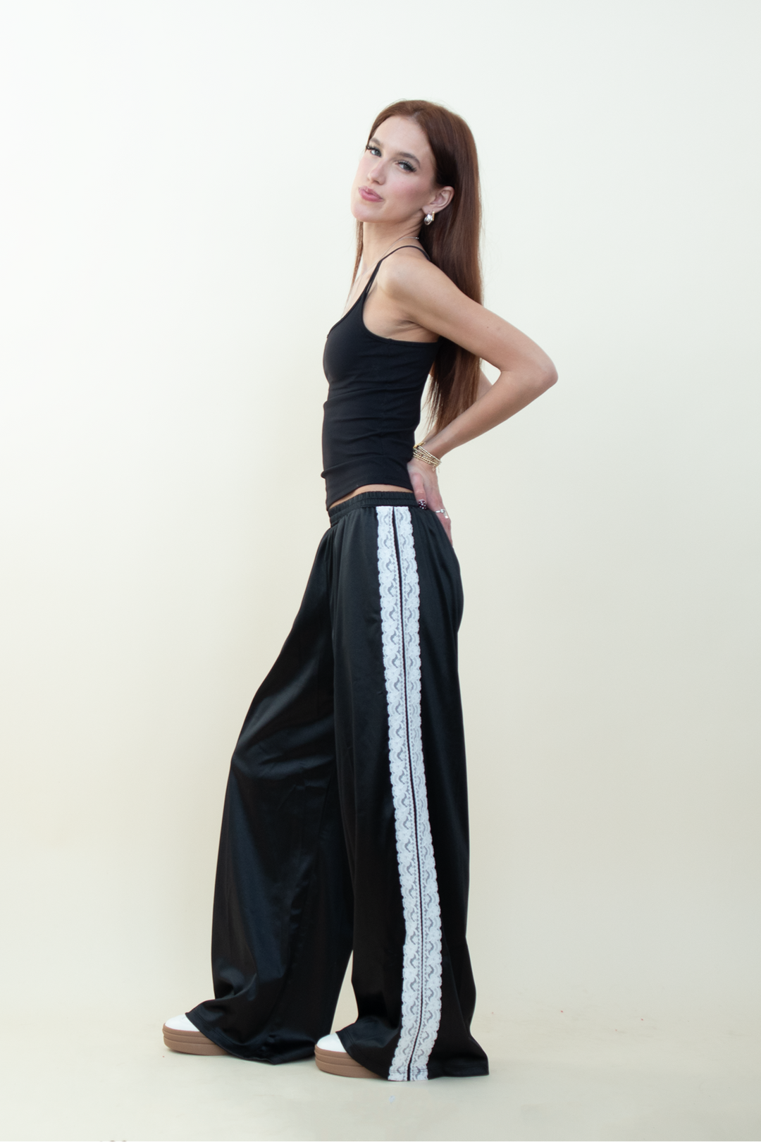 Model posing in black satin pants featuring white lace side panel, paired with a simple black tank top and platform sneakers.