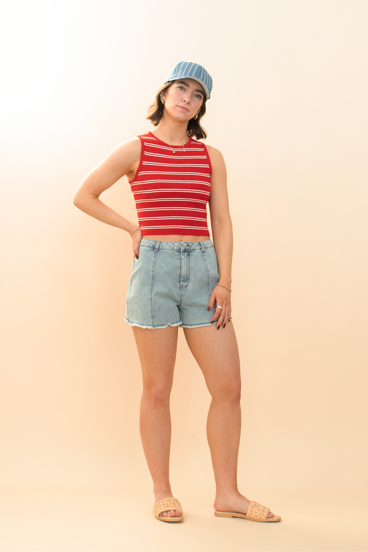 Trendy summer streetwear look: woman wearing striped tank top, high-waisted denim shorts with star patch, and blue pinstripe cap, showcasing a relaxed, Americana-inspired style.