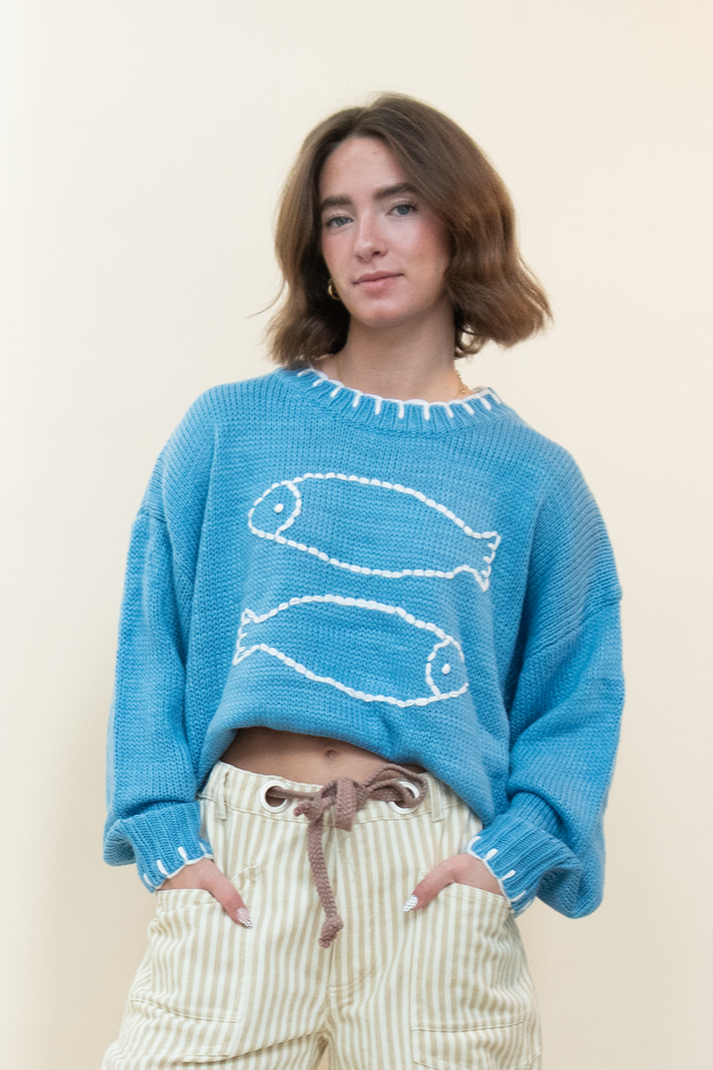 Woman wearing a blue fish sweater, styled with striped tan and white barrel jeans and light brown shoes. 