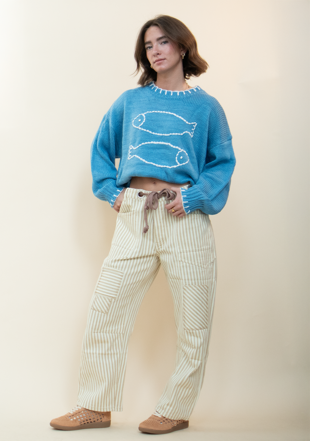 blue sweater with white fish stitched in the center and white stitch detailing around the neck and end of the sleeves, paired with pinstriped barrel jeans and tan sneakers.