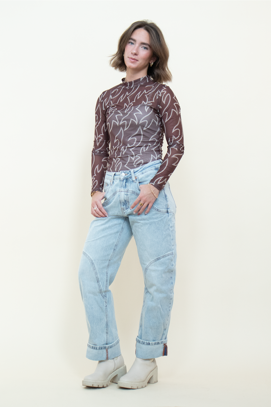 Model wearing a fitted brown mesh mock-neck top with cream Western-style print, styled with light-wash cuffed jeans.
