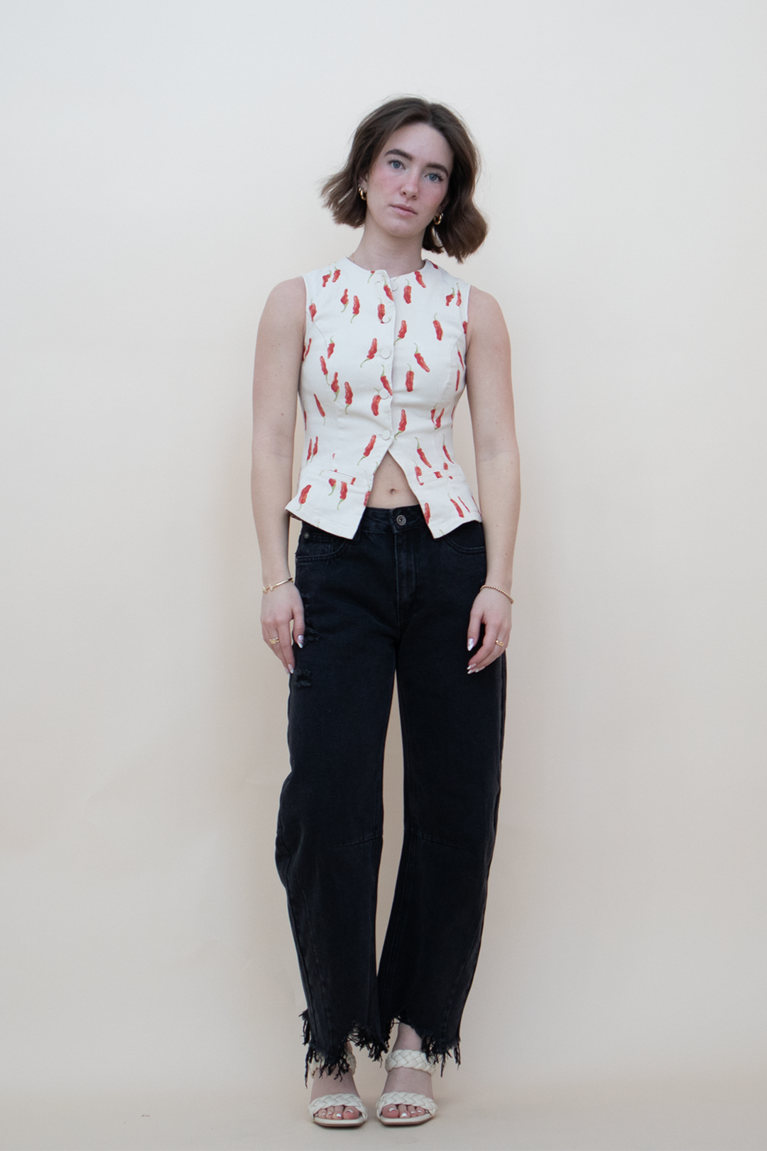 Woman wearing a sleeveless cream button-up vest top with red chili pepper print, paired with high-waisted black wide-leg jeans and white woven heeled sandals, standing against a neutral background — summer statement top for women.