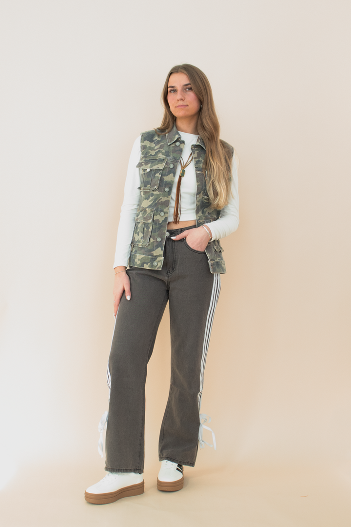 Full-body view of a woman wearing a sleeveless camo utility vest with flap pockets, layered over a white long sleeve crop top. She pairs it with high-waisted black athletic-style pants featuring side stripes and bow details near the hem, and white platform sneakers. Neutral background enhances the camo vest as the focal fashion piece.