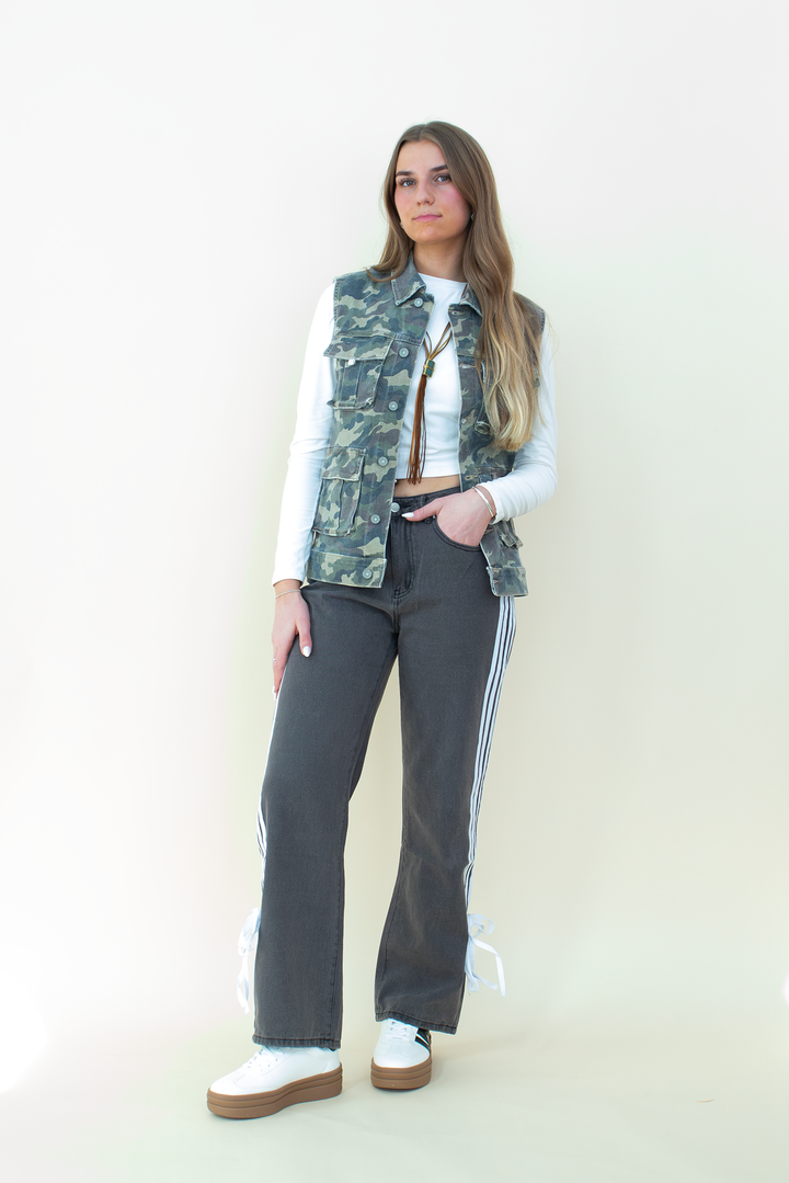 Full-body view of a woman wearing a sleeveless camo utility vest with flap pockets, layered over a white long sleeve crop top. She pairs it with high-waisted black athletic-style pants featuring side stripes and bow details near the hem, and white platform sneakers. Neutral background enhances the camo vest as the focal fashion piece.