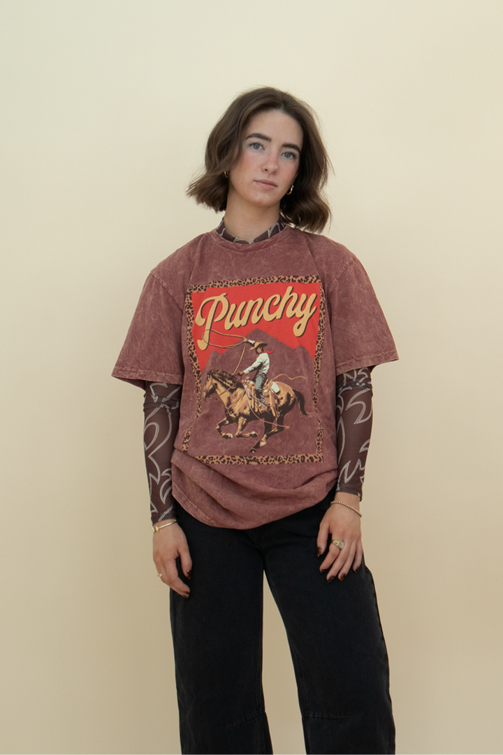 red-brown acid wash cowboy graphic tee with the phrase "punchy" printed on it, styled with a black jean and a mesh undershirt.