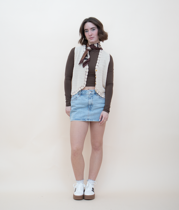 Cream crochet knit vest with gold buttons and lace trim, styled over a brown long-sleeve crop top with a denim mini skirt, neck scarf, and white sneakers