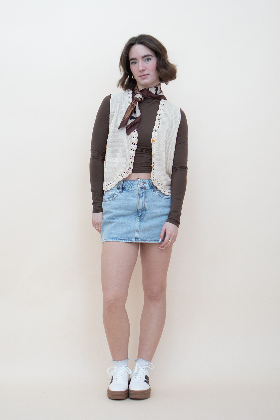 Cream crochet knit vest with gold buttons and lace trim, styled over a brown long-sleeve crop top with a denim mini skirt, neck scarf, and white sneakers