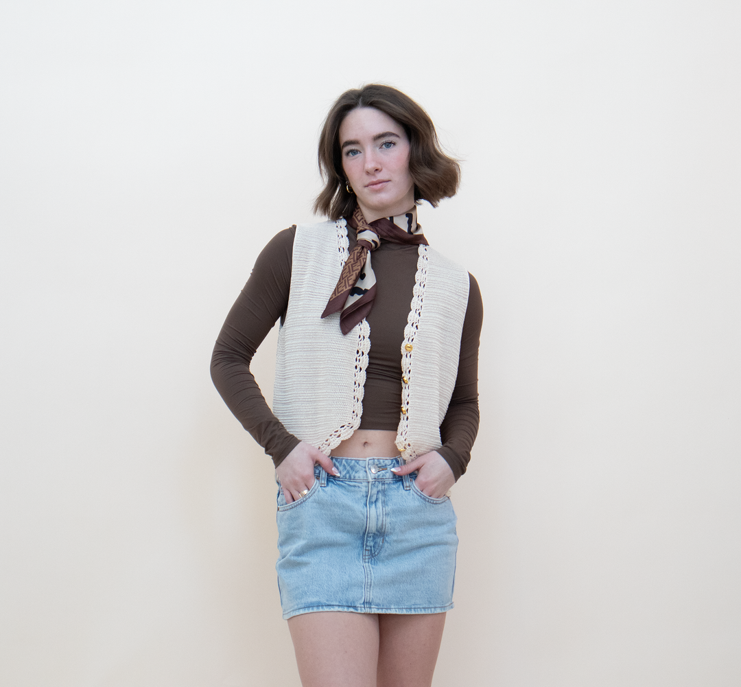 Close-up of a women's cream crochet knit vest showing gold button closure and delicate lace trim detail, styled with a brown long-sleeve crop top and a printed neck scarf