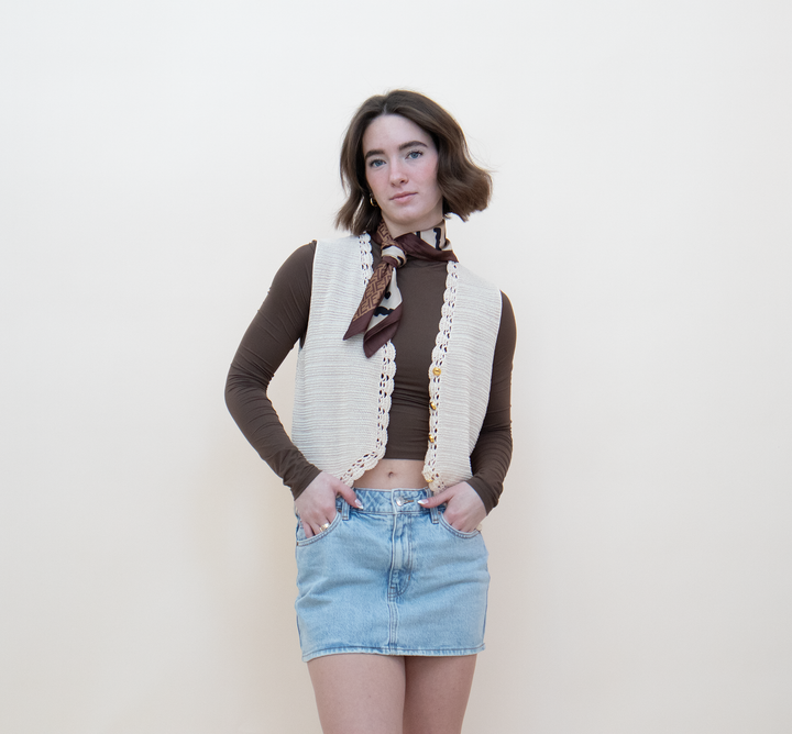Close-up of a women's cream crochet knit vest showing gold button closure and delicate lace trim detail, styled with a brown long-sleeve crop top and a printed neck scarf