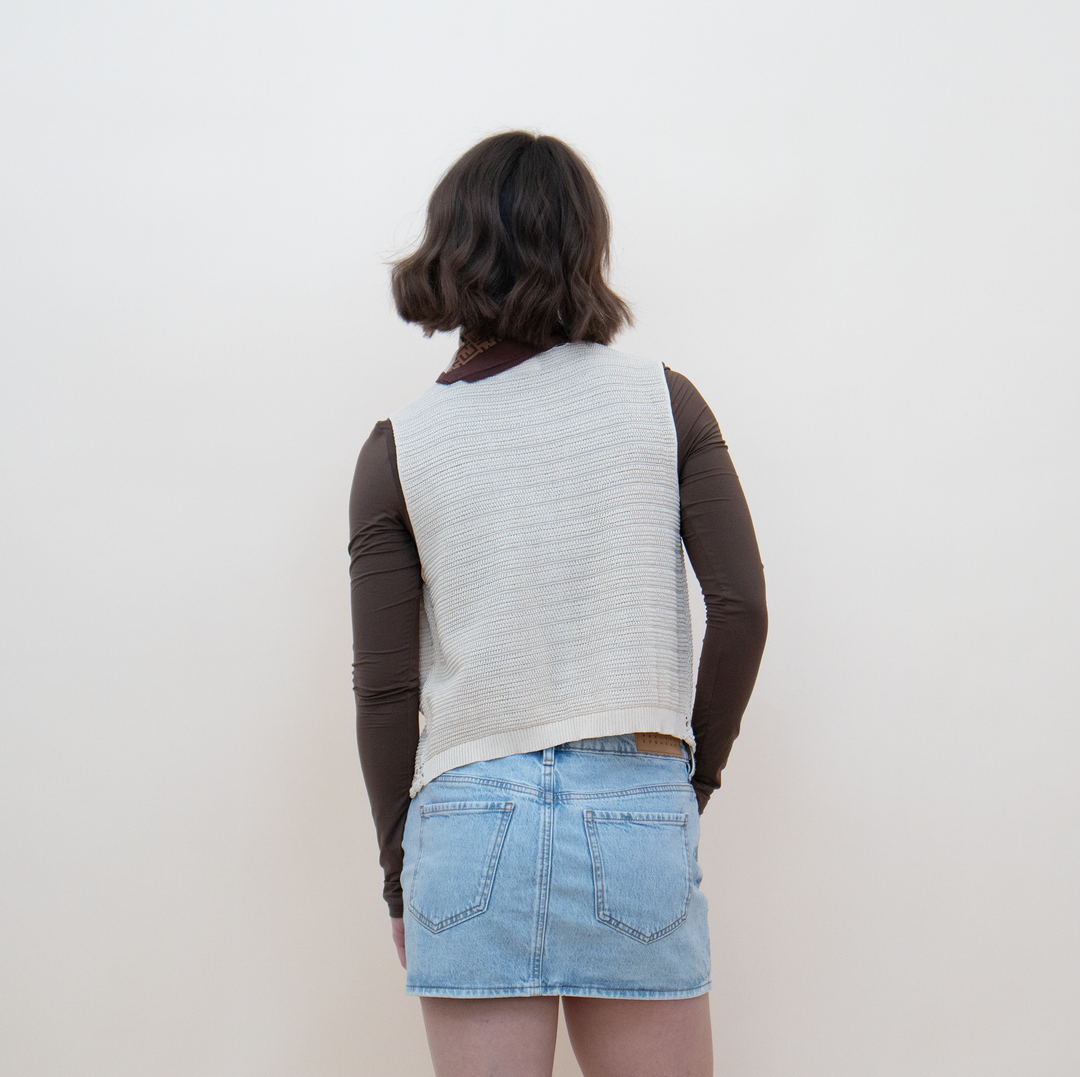 Close-up view of the back of a women's cream crochet knit vest with delicate lace trim detail, styled with a brown long-sleeve crop top and a mini skirt