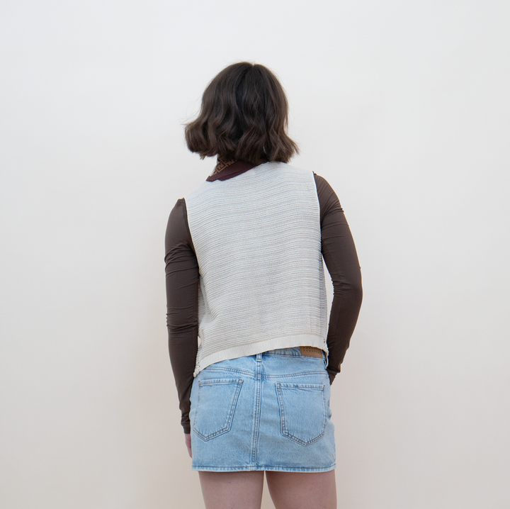 Close-up view of the back of a women's cream crochet knit vest with delicate lace trim detail, styled with a brown long-sleeve crop top and a mini skirt
