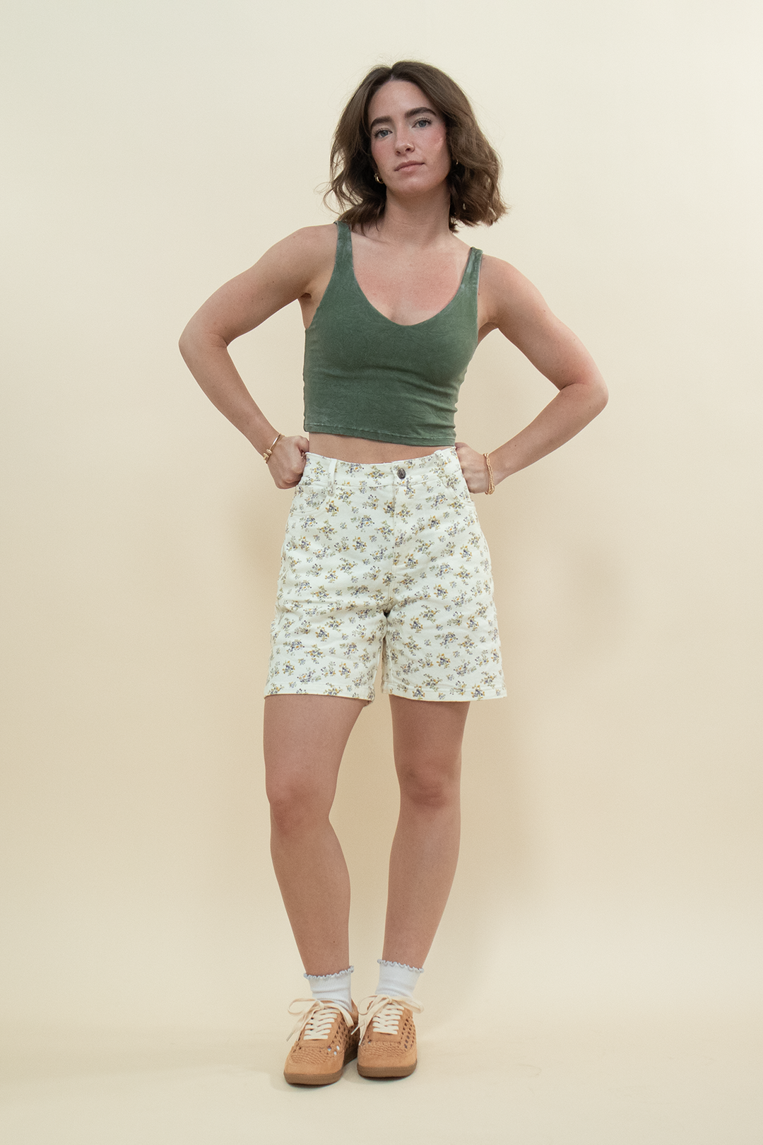 Model wearing a fitted green tank top and high-waisted floral denim shorts with tan sneakers, styled for a casual summer outfit idea.