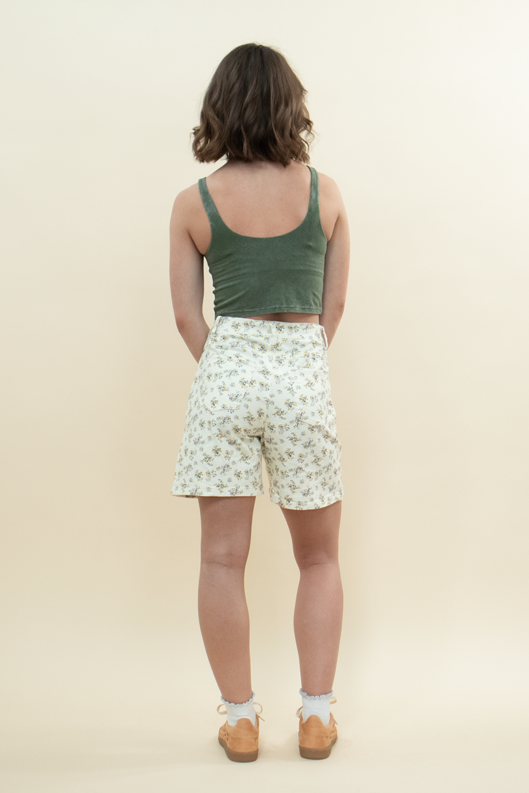 Back view of a woman wearing high-waisted floral print shorts paired with a fitted green crop tank top and tan sneakers, showcasing a casual summer look from behind.