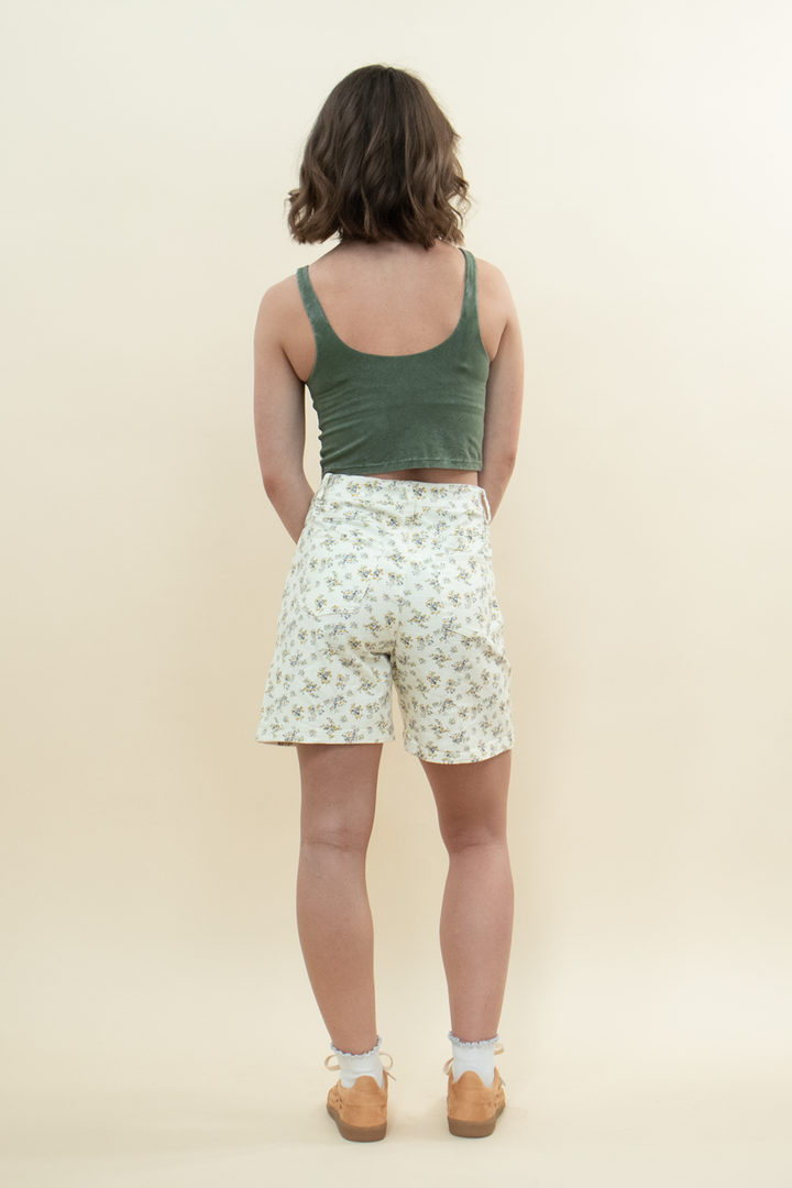 Back view of a woman wearing high-waisted floral print shorts paired with a fitted green crop tank top and tan sneakers, showcasing a casual summer look from behind.