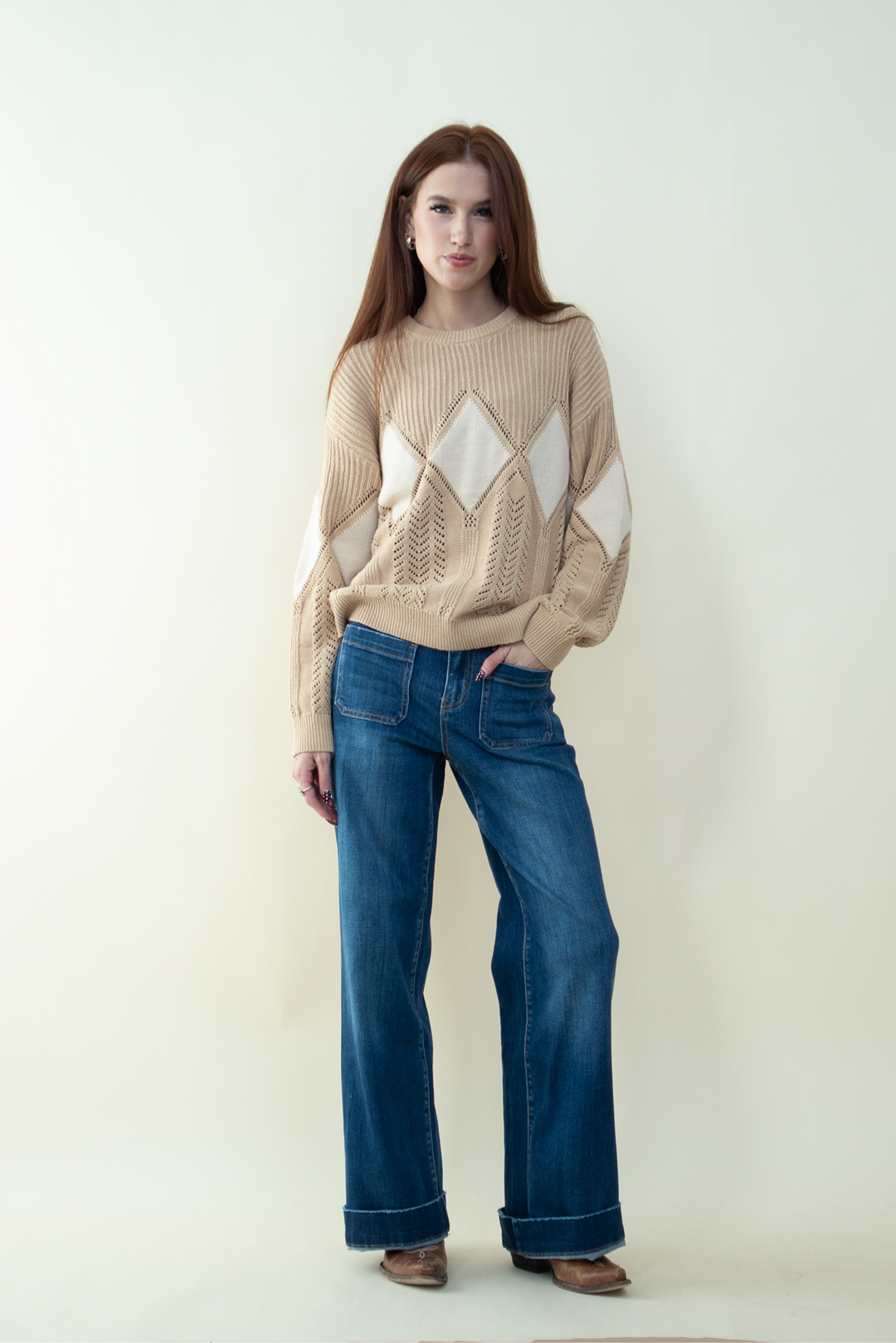 Woman wearing a beige knit sweater with diamond-shaped patterns and wide-leg blue jeans, standing against a light neutral background with one hand in her pocket.