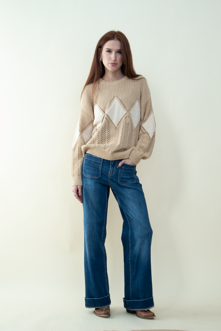 Woman wearing a beige knit sweater with diamond-shaped patterns and wide-leg blue jeans, standing against a light neutral background with one hand in her pocket.