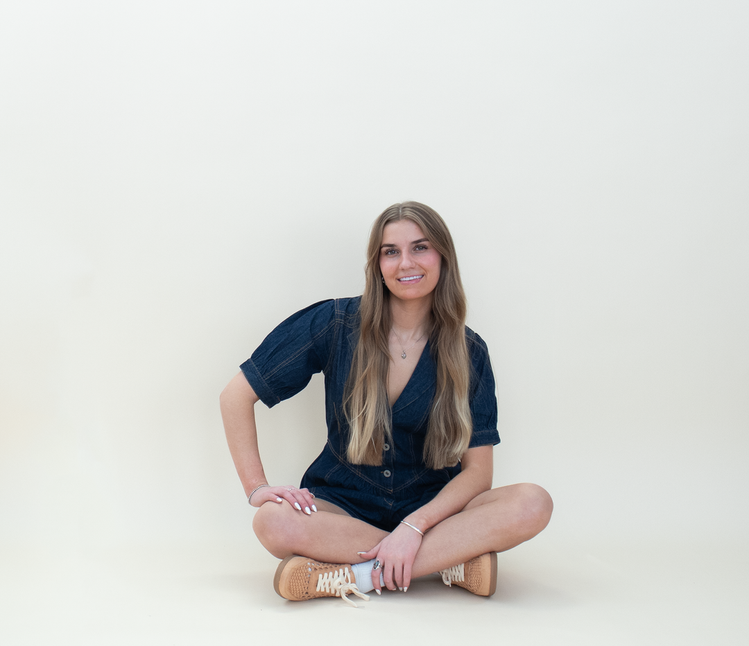 Women's dark wash denim puff sleeve romper with a deep V-neckline and button-front closure, shown in a relaxed seated pose with tan sneakers