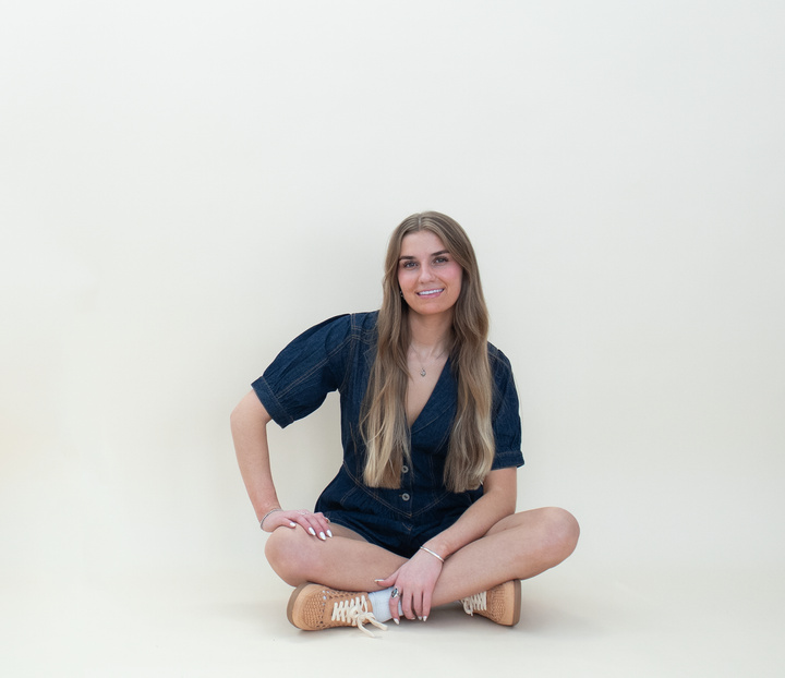 Women's dark wash denim puff sleeve romper with a deep V-neckline and button-front closure, shown in a relaxed seated pose with tan sneakers