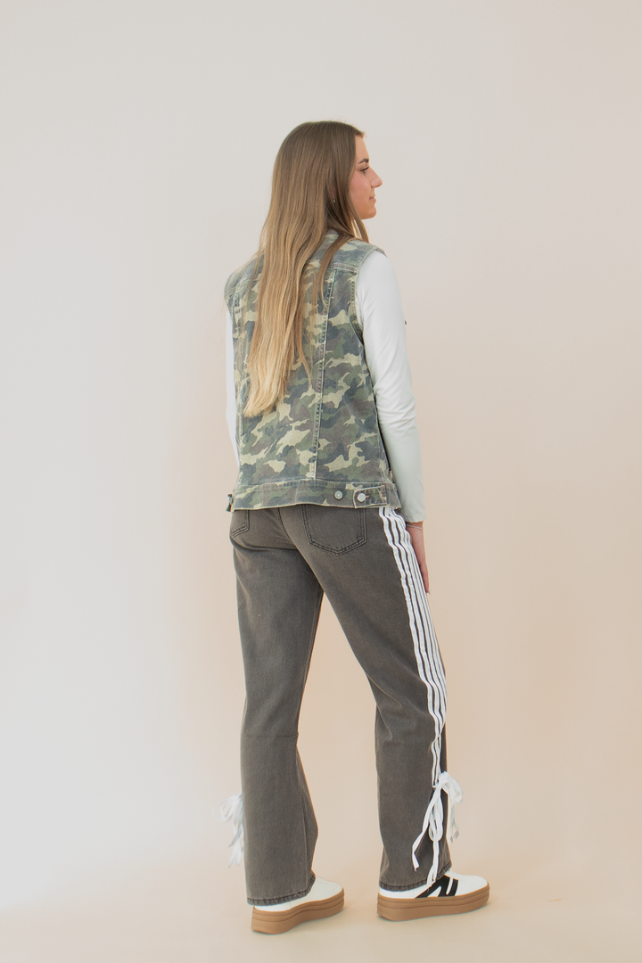 Back view of a woman wearing a sleeveless camouflage utility vest with flap pocket detailing, layered over a white long sleeve top. The camo print vest stands out against dark high-waisted pants with white side stripes and bow accents near the hem. Her long hair flows over the vest, completing the street-style inspired look.