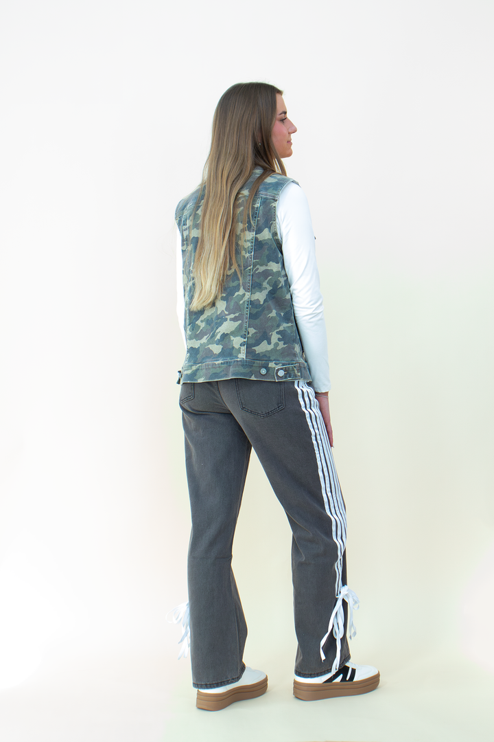 Back view of a woman wearing a sleeveless camouflage utility vest with flap pocket detailing, layered over a white long sleeve top. The camo print vest stands out against dark high-waisted pants with white side stripes and bow accents near the hem. Her long hair flows over the vest, completing the street-style inspired look.
