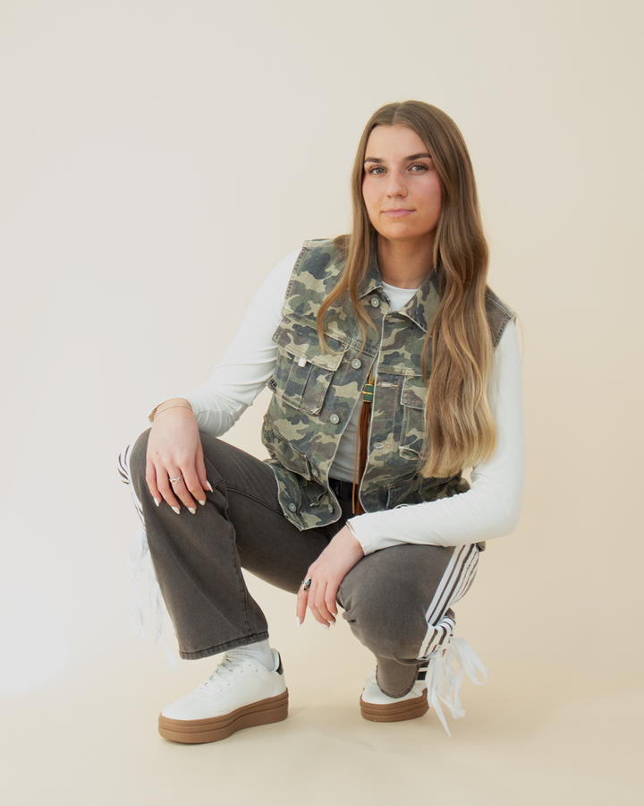 View of a crouched woman wearing a sleeveless camo utility vest with flap pockets, layered over a white long sleeve crop top. She pairs it with high-waisted black athletic-style pants featuring side stripes and bow details near the hem, and white platform sneakers. Neutral background enhances the camo vest as the focal fashion piece.