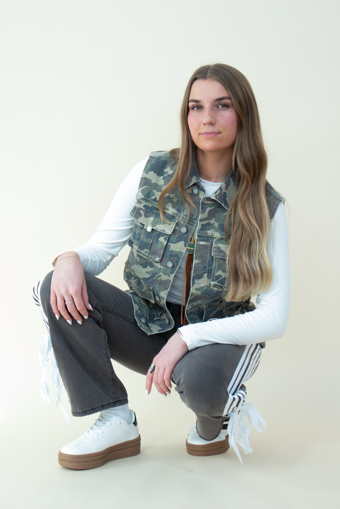 View of a crouched woman wearing a sleeveless camo utility vest with flap pockets, layered over a white long sleeve crop top. She pairs it with high-waisted black athletic-style pants featuring side stripes and bow details near the hem, and white platform sneakers. Neutral background enhances the camo vest as the focal fashion piece.