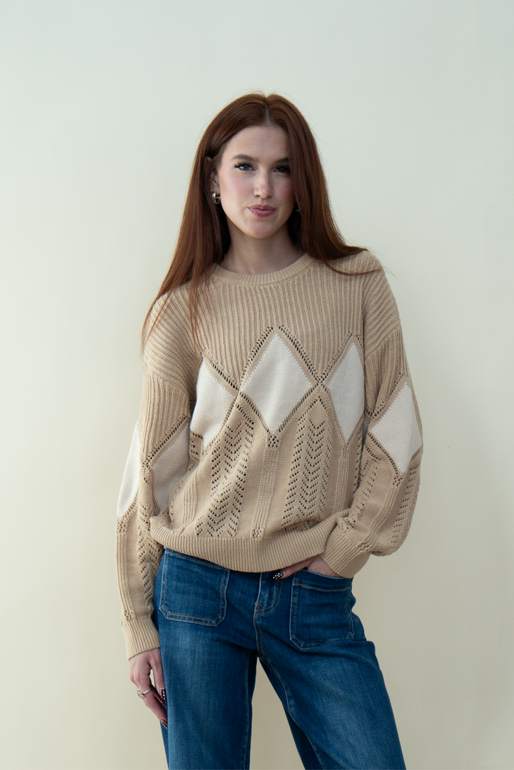 Woman with long straight auburn hair wearing a beige knit sweater with large white diamond patterns, standing against a light background.