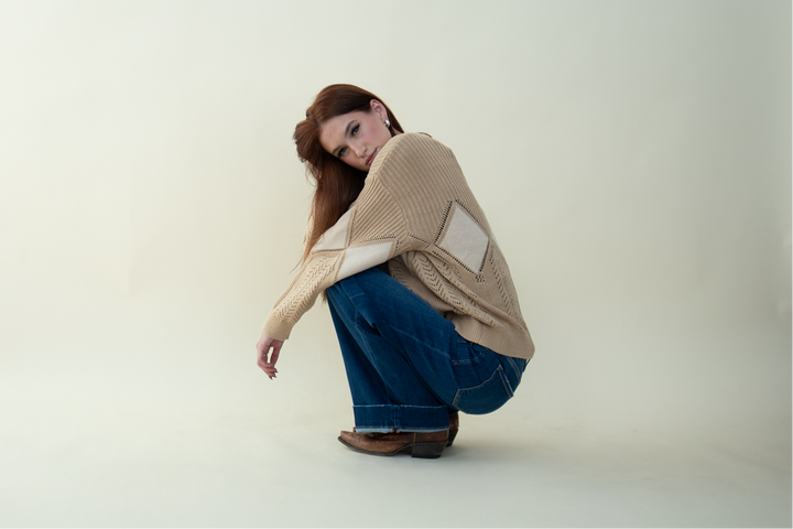 Model wearing a beige knit sweater with diamond cut-out details, wide-leg blue jeans, and brown boots, posing in a crouched position.