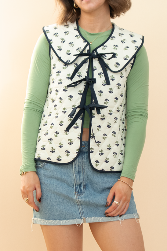 Woman modeling a floral quilted vest with oversized collar and navy bow ties, styled with a light green long sleeve top and raw hem denim mini skirt – feminine layered outfit for spring fashion.