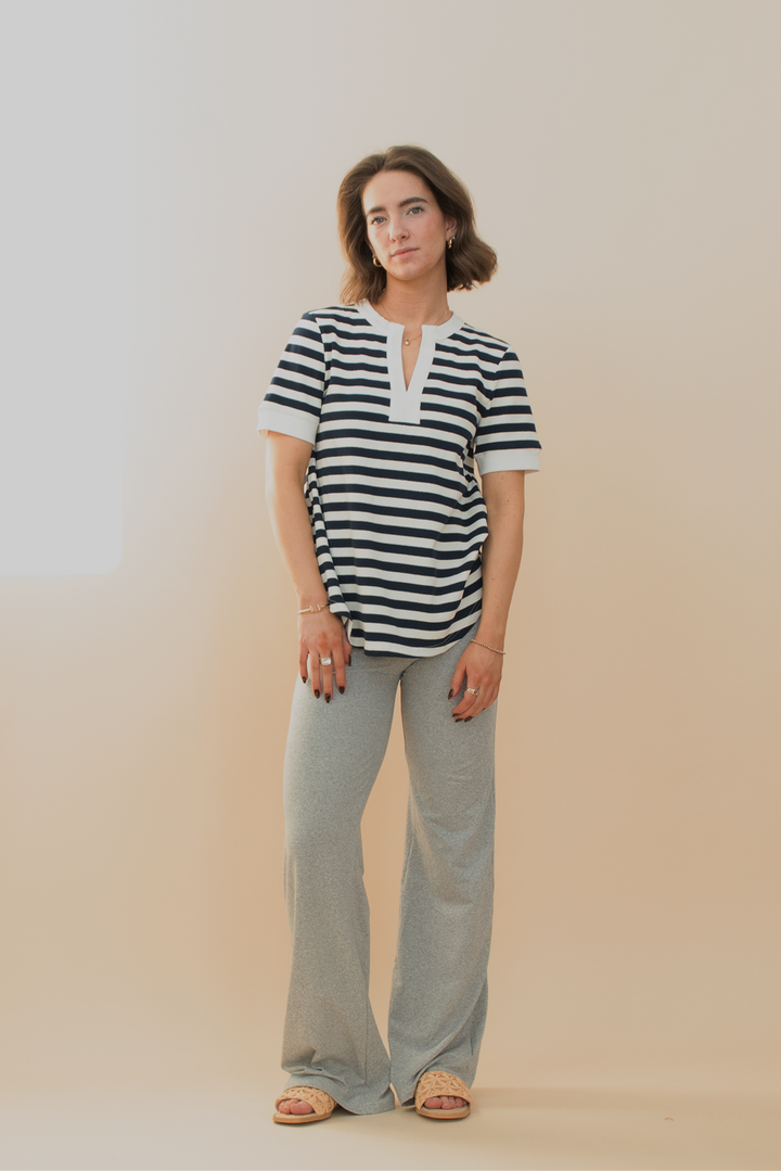 Woman wearing a navy and white striped short-sleeve top with a split neckline, paired with light gray lounge flare pants and woven slide sandals, posing against a soft beige background.